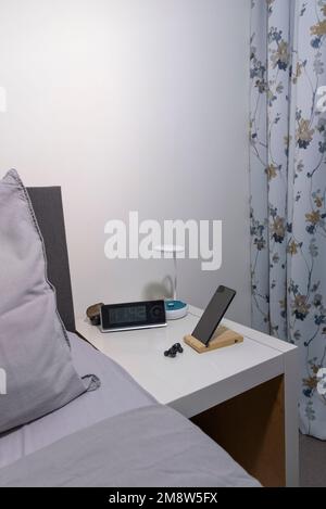 Smartphone on the bedside table with lamp, headset, clock and fragment of bed. Vertical view Stock Photo