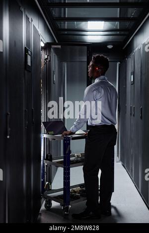 Vertical full length portrait of black man as network engineer using laptop and setting up servers in data center Stock Photo