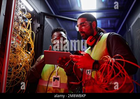 Waist up portrait of two technicians using digital tablet while repairing servers and setting up data network in neon light Stock Photo
