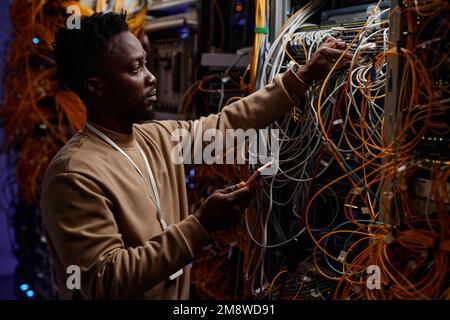 Side view portrait of black young man as system administrator fixing server with messy cables and wires Stock Photo