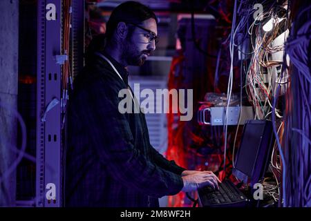 Side view portrait of bearded system administrator setting up server network and using laptop in neon light Stock Photo