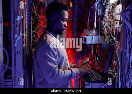 Side view portrait of black man as network administrator setting up servers and using laptop in neon light Stock Photo