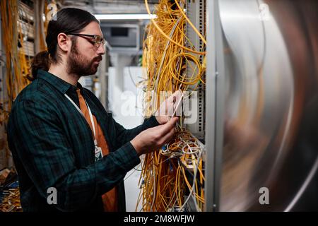 Side view portrait of bearded system administrator setting up server and using smartphone Stock Photo