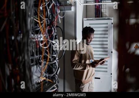 Moody wide angle shot of black man as system administrator standing in server room and holding tablet, copy space Stock Photo