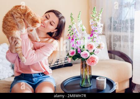 Playing with ginger cat at home by fresh bouquet. Young woman relaxing on couch in living room and holding pet. Feeling cozy with kitten and having fu Stock Photo