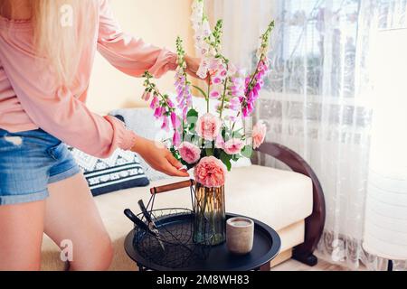 Woman puts bouquet of roses and foxgloves flowers picked in basket in vase at home. Interior and summer decor on coffee table. Fresh pink purple orang Stock Photo