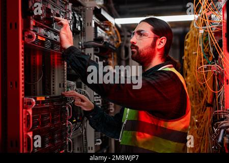 Side view portrait of bearded network technician inspecting servers and doing maintenance work in data center lit by red neon light Stock Photo