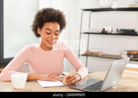 Cheerful pretty young mixed race curly woman manager making notes at table with laptop, watch online lesson Stock Photo