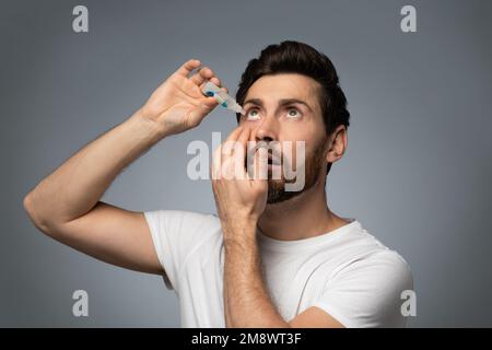 Handsone bearded caucasian man applying ophthalmology eyedropper, standing on grey studio background Stock Photo