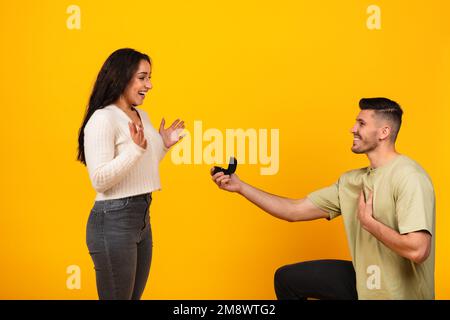Smiling millennial arab guy kneeling giving ring box to happy excited bride, isolated on orange background Stock Photo