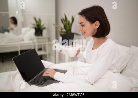 Positive pretty woman using laptop in bed, mockup Stock Photo
