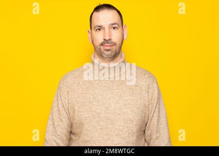 Handsome bearded hispanic man wearing beige turtleneck sweater posing smiling for studio portrait over yellow background. Stock Photo