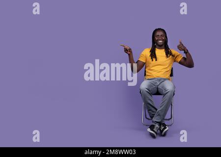 Happy black guy sitting on chair, pointing at copy space Stock Photo