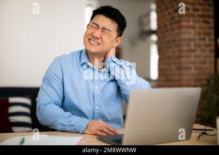 Neck ache. Mature asian man massaging aching neck, suffering from pain while working on laptop at table at home office Stock Photo