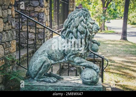 Lion sculpture with paw on ball - one of two beside estate entrancegate with leafy  Neighborhood street blurred and washed out in background Stock Photo