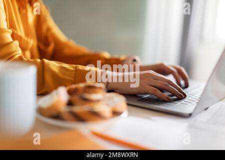 Female Hands Typing On Laptop Keyboard While Sitting At Table In Kitchen Stock Photo
