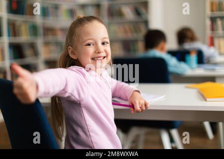 Happy caucasian schoolgirl gesturing thumbs up and smiling to camera, sitting at school in classroom, free space Stock Photo