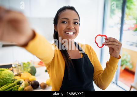 Smiling young african american female in apron with piece of pepper makes selfie with vegetables Stock Photo