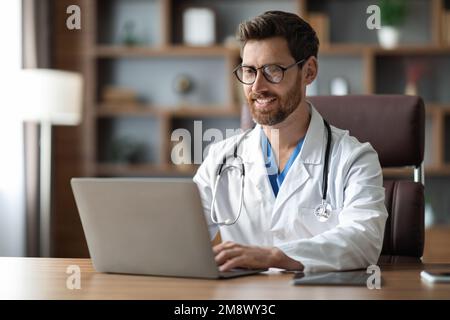 Smiling Male Doctor Working On Laptop While Sitting At Desk In Office Stock Photo