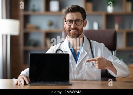 Doctor pointing at blank laptop screen while sitting at desk in office Stock Photo