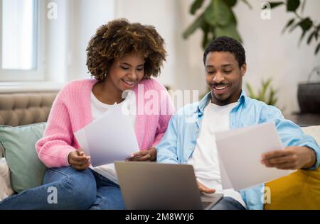 Positive black couple reading documentation at home, using laptop Stock Photo