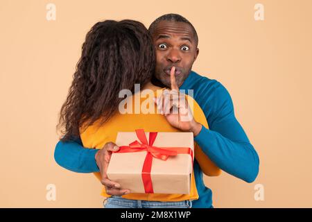 Loving black middle aged husband hugging wife holding gift behind her back and gesturing hush sign, preparing surprise Stock Photo