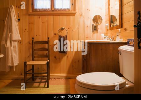 Antique wooden high back chair, vanity and toilet in main bathroom on upstairs floor inside milled log home. Stock Photo