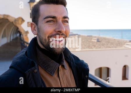 Ethnic bearded man smiling and looking away on top of old building located near sea in daylight Stock Photo