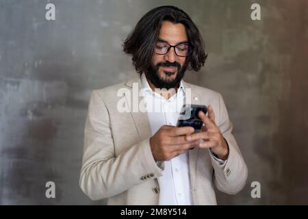 A young Arab businessman in shirt and beige jacket with long hair and beard, smiles using a smartphone in a neutral office setting with an antiqued wa Stock Photo