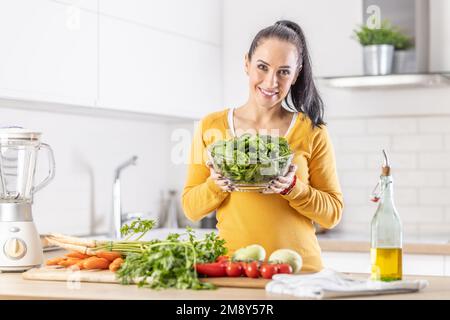 Smiling woman with a bowl full of spinach leaves along with fresh vegetables and olive oil in her kitchen. Stock Photo