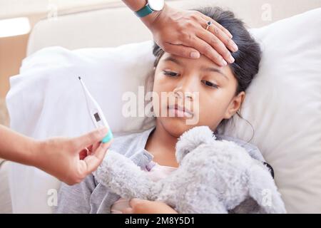 Little girl having her temperature taken with a thermometer by her mother at home. Small sick child lying in bed holding her toy, while a doctor Stock Photo