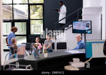 Practitioner nurse bringing elderly patient to family after finishing medical consultation, granddaughter holding bouquet flowers for grandmother. People standing in hospital reception. Stock Photo