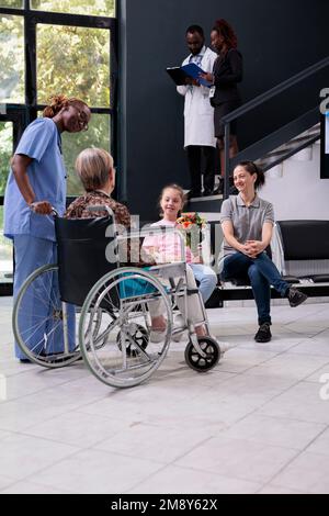 Medical assistant bringing old patient to family after finishing appointment while explaining health care treatment in hospital waiting area. Granddaughter holding bouquet of flowers for grandmother Stock Photo