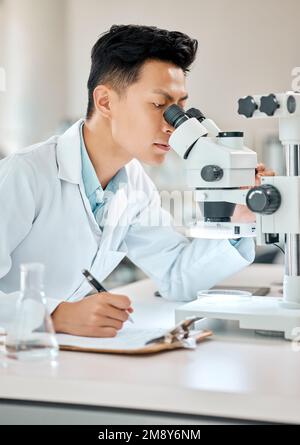 I wont let anything escape my eye. a young scientist writing notes while using a microscope in a lab. Stock Photo