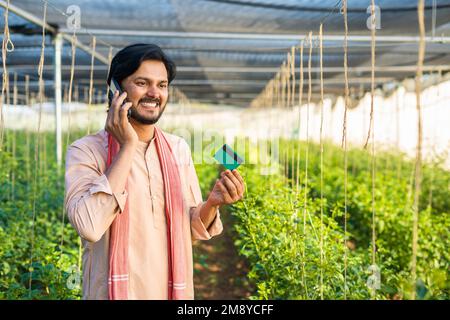 young farmer at greenhouse talking on mobile phone call by holding green credit card - conept of online payment, customer service and agronomy Stock Photo