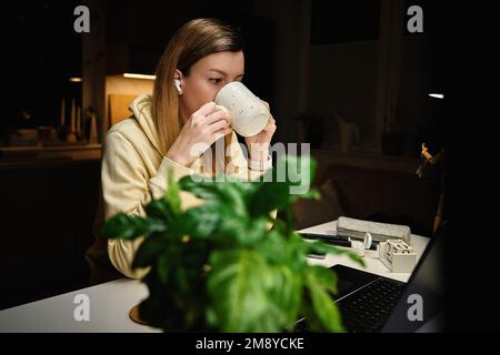 Focused woman drinks coffee and looking at laptop screen. Female freelancer works in home office workplace. Overtime work at night Stock Photo