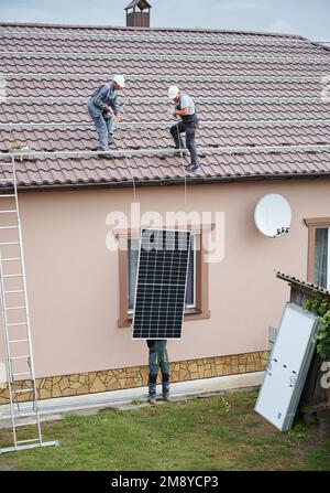 Men technicians lifting up photovoltaic solar modul on roof of house. Engineers in helmets building solar panel system outdoors. Concept of alternative and renewable energy. Stock Photo