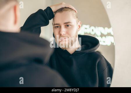 An attractive young man with a beard looks at himself in the mirror. Stock Photo