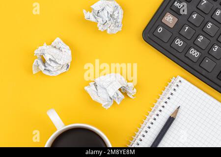 Notepad, calculator, coffee cup and crumpled paper balls on a yellow background. Stock Photo