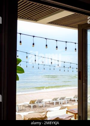 Hanging festive string wired light bulbs decorate under the roof outside the cafe on beachfront with seascape view background, vertical style. Stock Photo