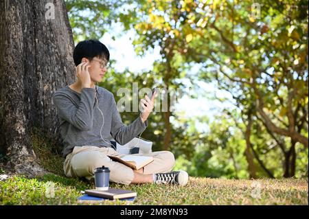 Happy and relaxed young Asian male college student listening to music while relaxing under the tree in the campus park. Stock Photo