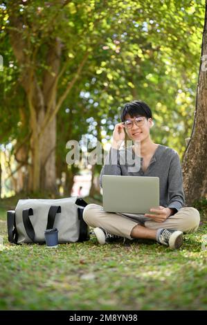 Portrait, Smart and happy young Asian male college student wearing glasses, sits under the tree in the campus park with his portable laptop computer. Stock Photo