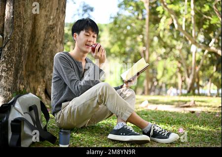 Relaxed and happy young Asian male college student eating apple while reading a book under the tree in the park. leisure concept Stock Photo