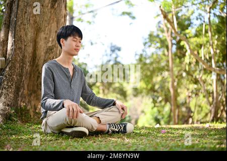 Handsome and relaxed young Asian man in casual clothes meditating under the tree in the greenery park. Stock Photo