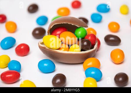 Half of a cracked chocolate egg filled with multi-colored candies covered with an egg shell on a white background with multi-colored candies. Treats f Stock Photo