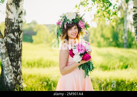 portrait of a young Caucasian girl with a wreath on her head and a bouquet of peonies in a dress outdoors in summer Stock Photo