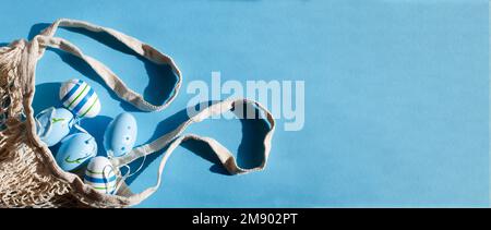 colorful handmade painted easter eggs in a string bag on blue background with hard shadows. minimal concept. Top view. An Easter card with a copy spac Stock Photo