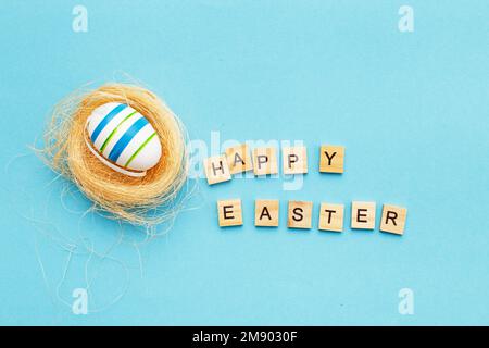 inscription happy easter with colorful handmade painted egg in the nest on blue background with copy space. Stock Photo