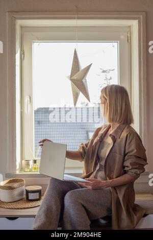 Woman working at home with laptop sitting on table near window. Stock Photo