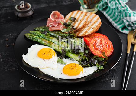 Breakfast. Fried egg, bread toast, green asparagus, tomatoes and jamon on black plate. Stock Photo
